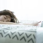2026 如何戒掉拖延症實測：一位年輕女性在清晨床鋪上試圖伸手關掉鬧鐘，展現大腦在前額葉皮質理智與邊緣系統逃避本能相互博弈下的日常啟動障礙場景。