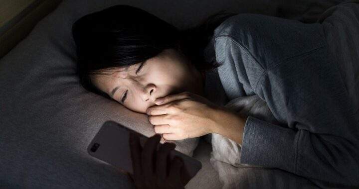 2026 擺脫熬夜拖延實測：在深夜漆黑的臥室中，一位夜貓子躺在床上被手機螢幕藍光照亮面部，展現現代人因白天壓力引發深夜報復性分心與睡眠拖延的真實生活痛點場景。