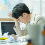 2026 高效時間管理術實測：一位辦公室上班族因為數位通知干擾與時間碎片化而焦慮扶額，桌面上散落著工作廢紙，展現現代人渴望透過自律微習慣克服拖延症的日常痛點場景。
